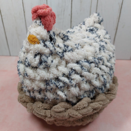 Handmade crochet chicken plush made with fluffy white and blue speckled polyester yarn, featuring a red comb, yellow beak, and sitting in a soft brown crocheted nest.